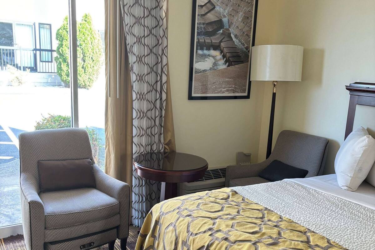 A hotel room with a bed, two armchairs, a small round table, a lamp, and a window with curtains.