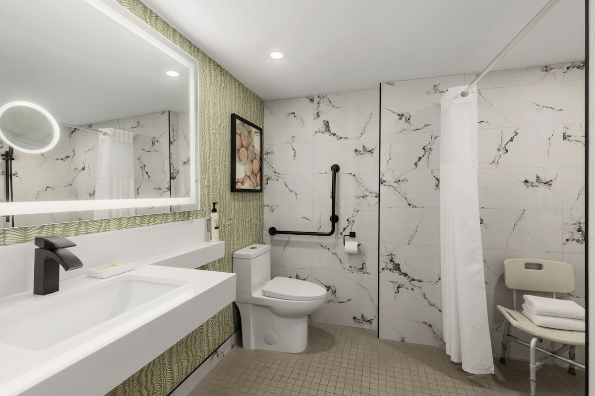 Une salle de bain moderne avec un lavabo, des toilettes et une douche blancs, des murs à motifs de marbre et un miroir avec éclairage intégré.