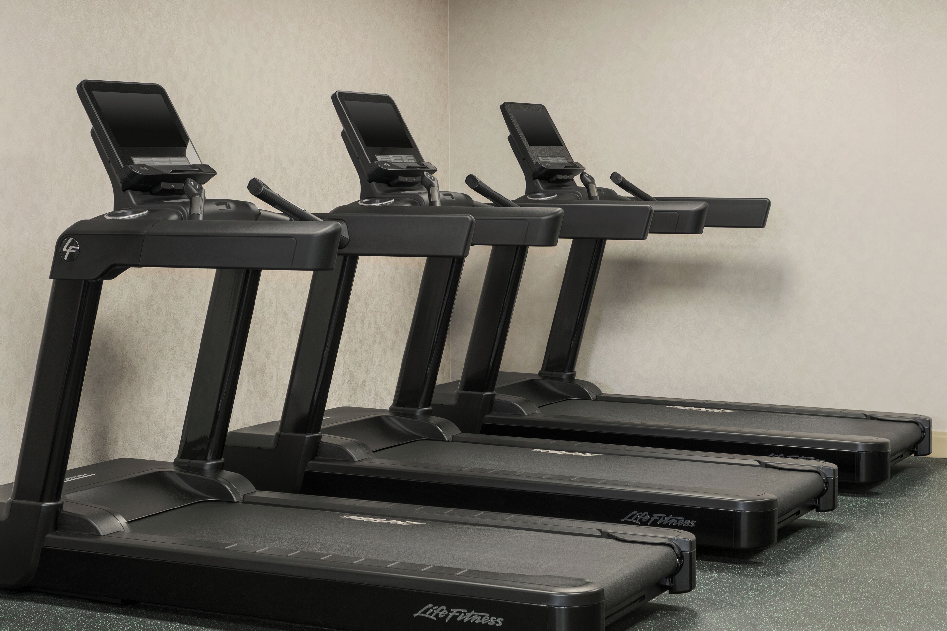 Four treadmills with control panels, arranged in a row against a plain wall.