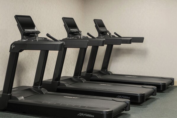 Four treadmills with control panels, arranged in a row against a plain wall.