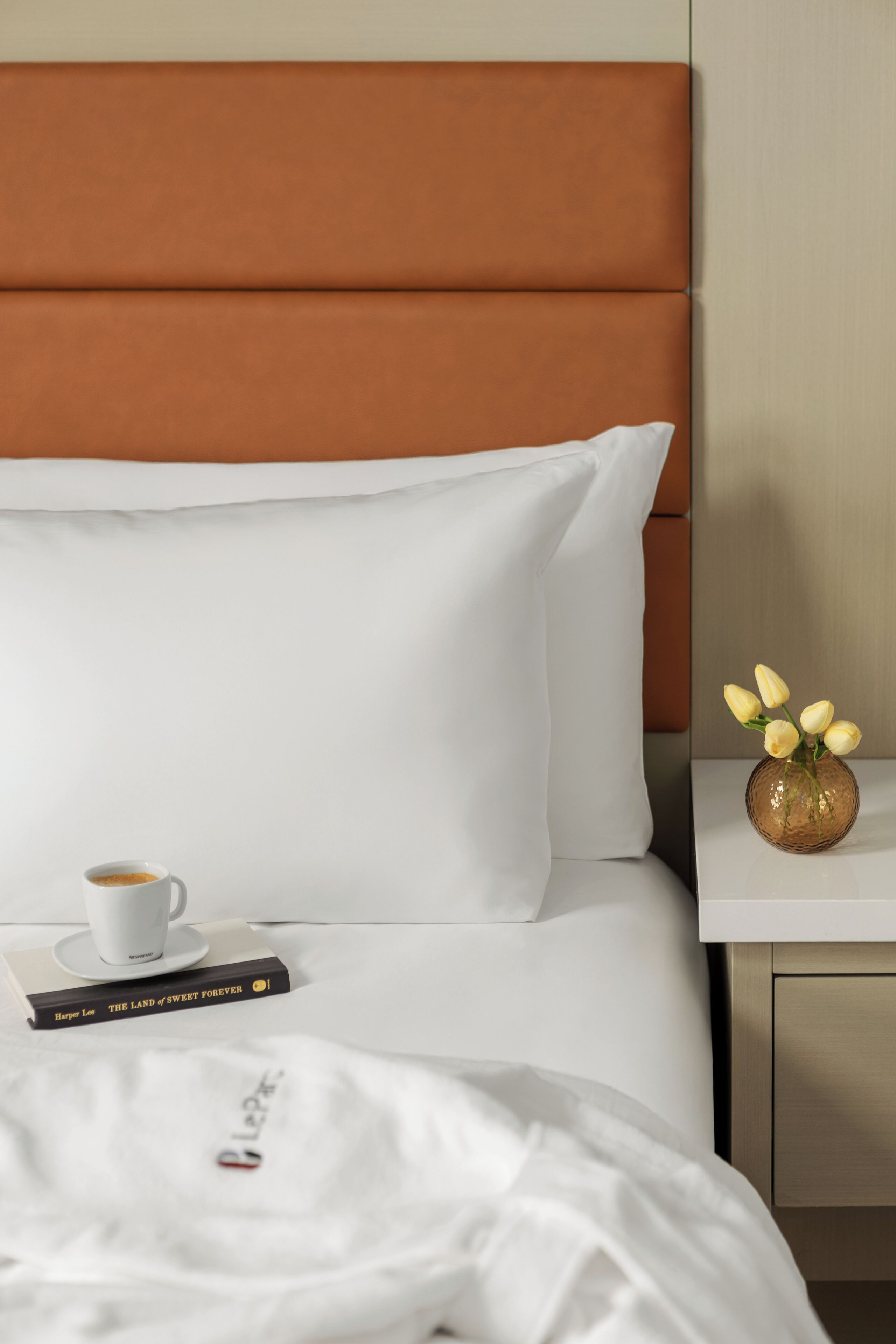 A neatly made bed with white linens, a brown headboard, a white pillow, a white cup, a book titled 'The Land of Sweet Forever,' and a small vase with yellow flowers on a bedside table.