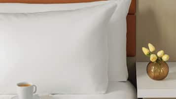 A neatly made bed with white linens, a brown headboard, a white pillow, a white cup, a book titled 'The Land of Sweet Forever,' and a small vase with yellow flowers on a bedside table.