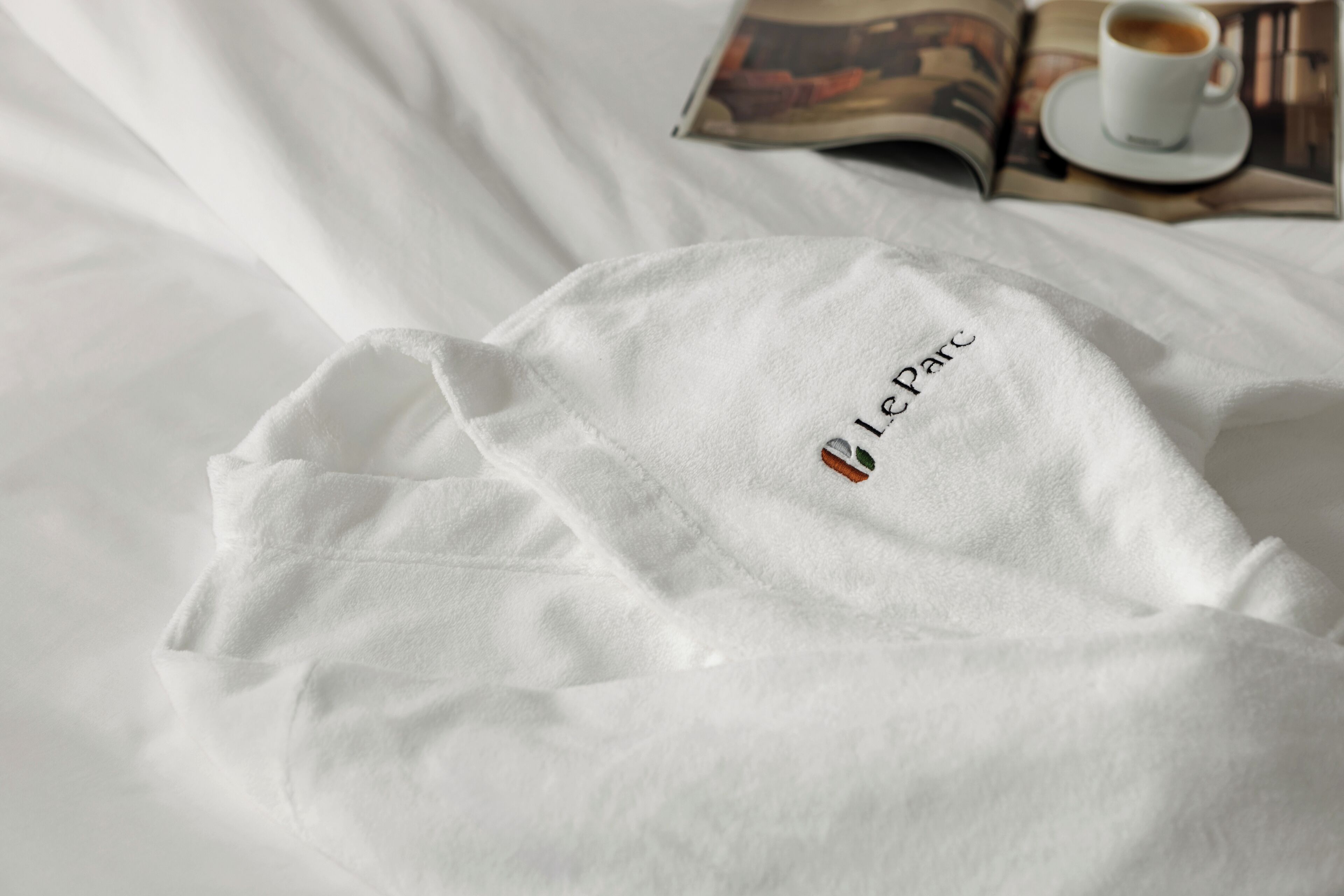 A white towel with 'Le Parc' embroidered on it, placed on a bed with a magazine and a cup of coffee in the background.