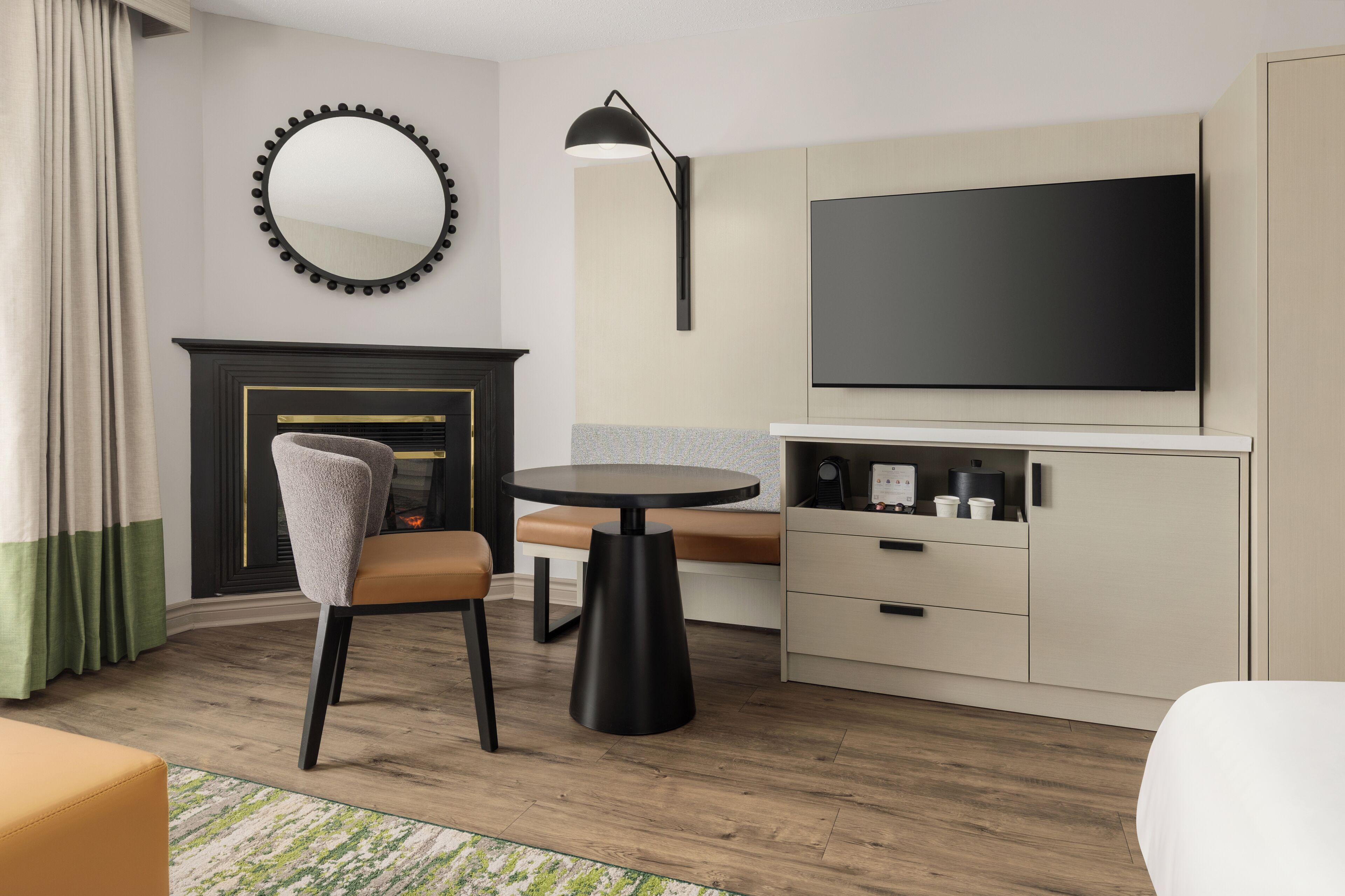 A modern hotel room with a round table, a sofa, a TV, a fireplace, and a cabinet with drawers.