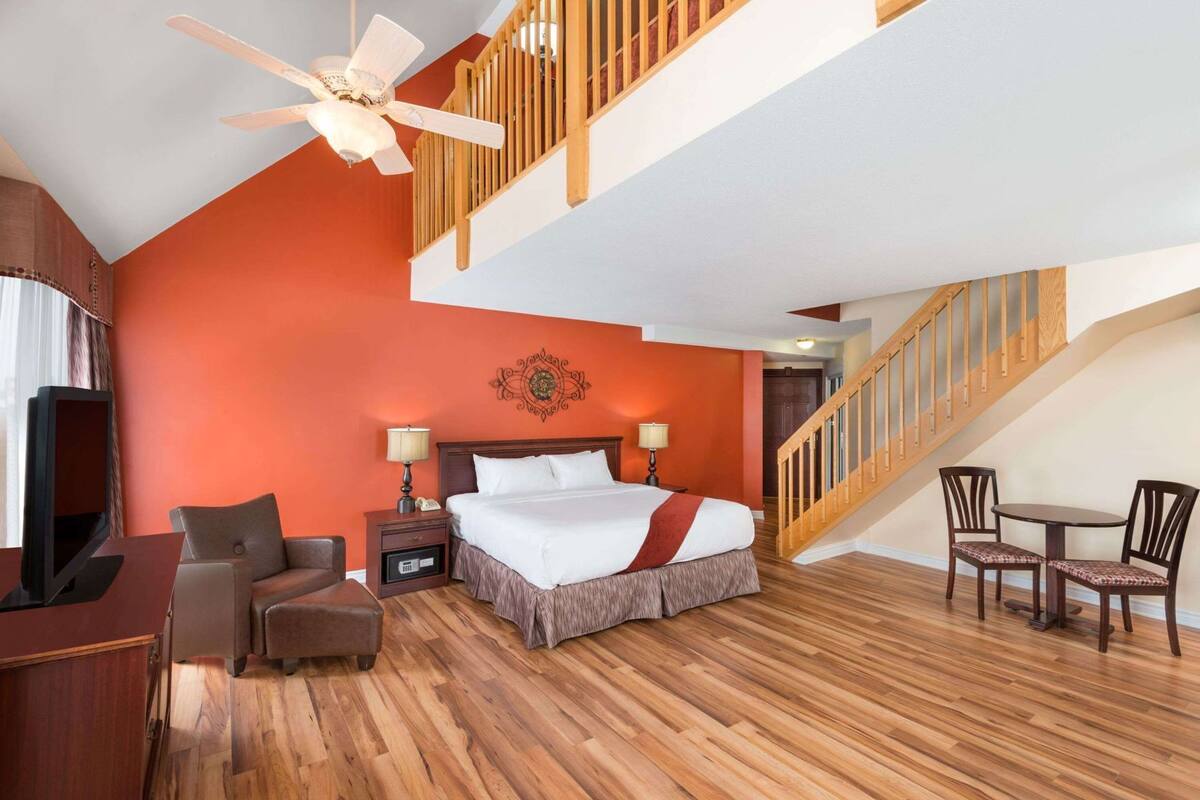 Une chambre d’hôtel comprenant un lit, un bureau avec un ordinateur, une télévision, une chaise, une table avec des chaises, un escalier et un ventilateur de plafond.