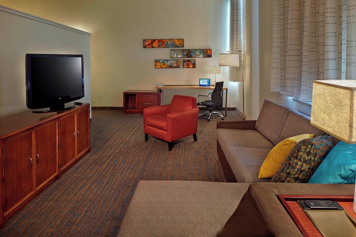 A hotel room with a television, a desk with a computer, a red armchair, a brown sofa with colorful pillows, and a wooden cabinet.