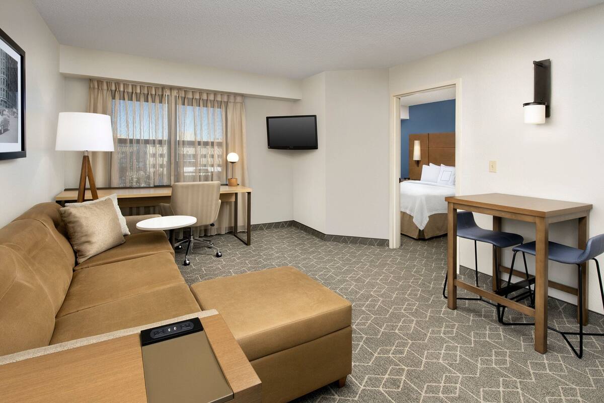 Una habitació d'hotel amb un sofà, una taula petita amb una làmpada, un televisor penjat a la paret i un llit a l'habitació propera.