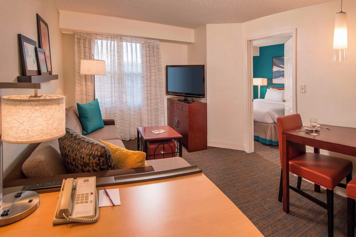 Une chambre d’hôtel comprenant un lit, un bureau avec un téléphone, une télévision, une table à manger avec des chaises et une fenêtre avec des rideaux.