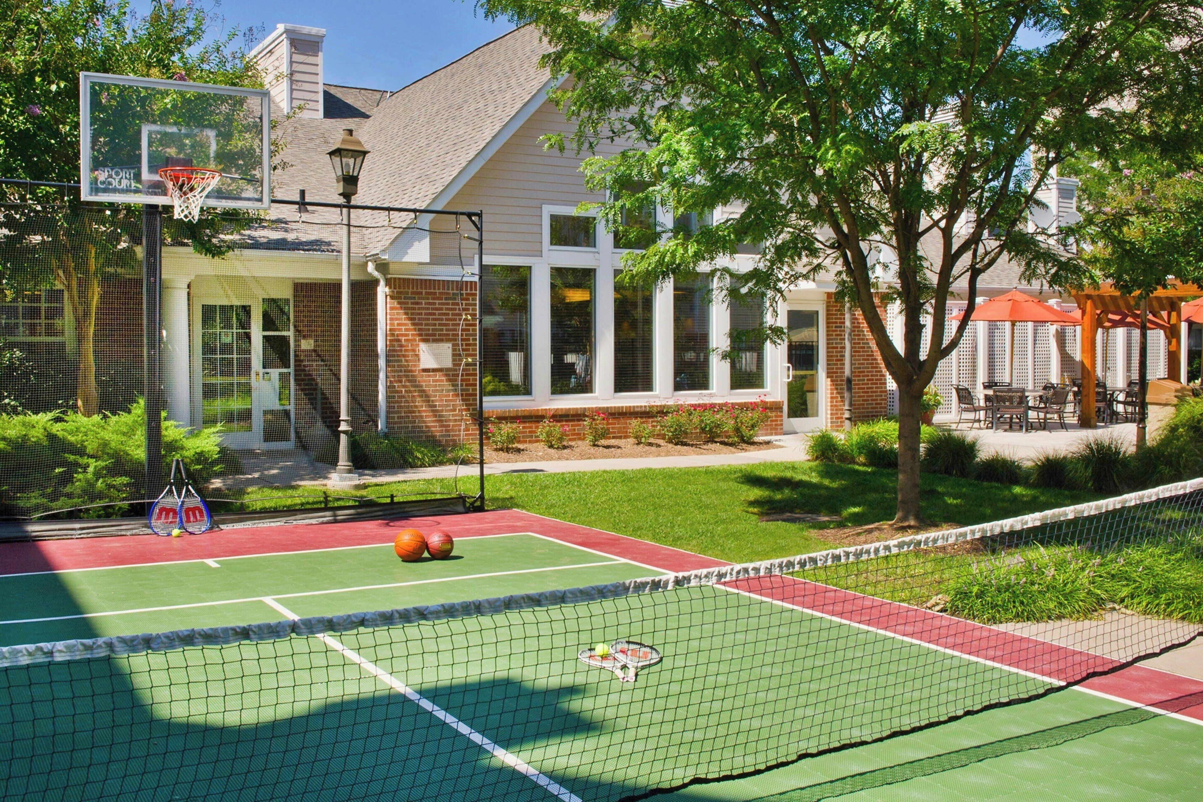 An outdoor tennis court with a net, a basketball hoop, and a basketball.