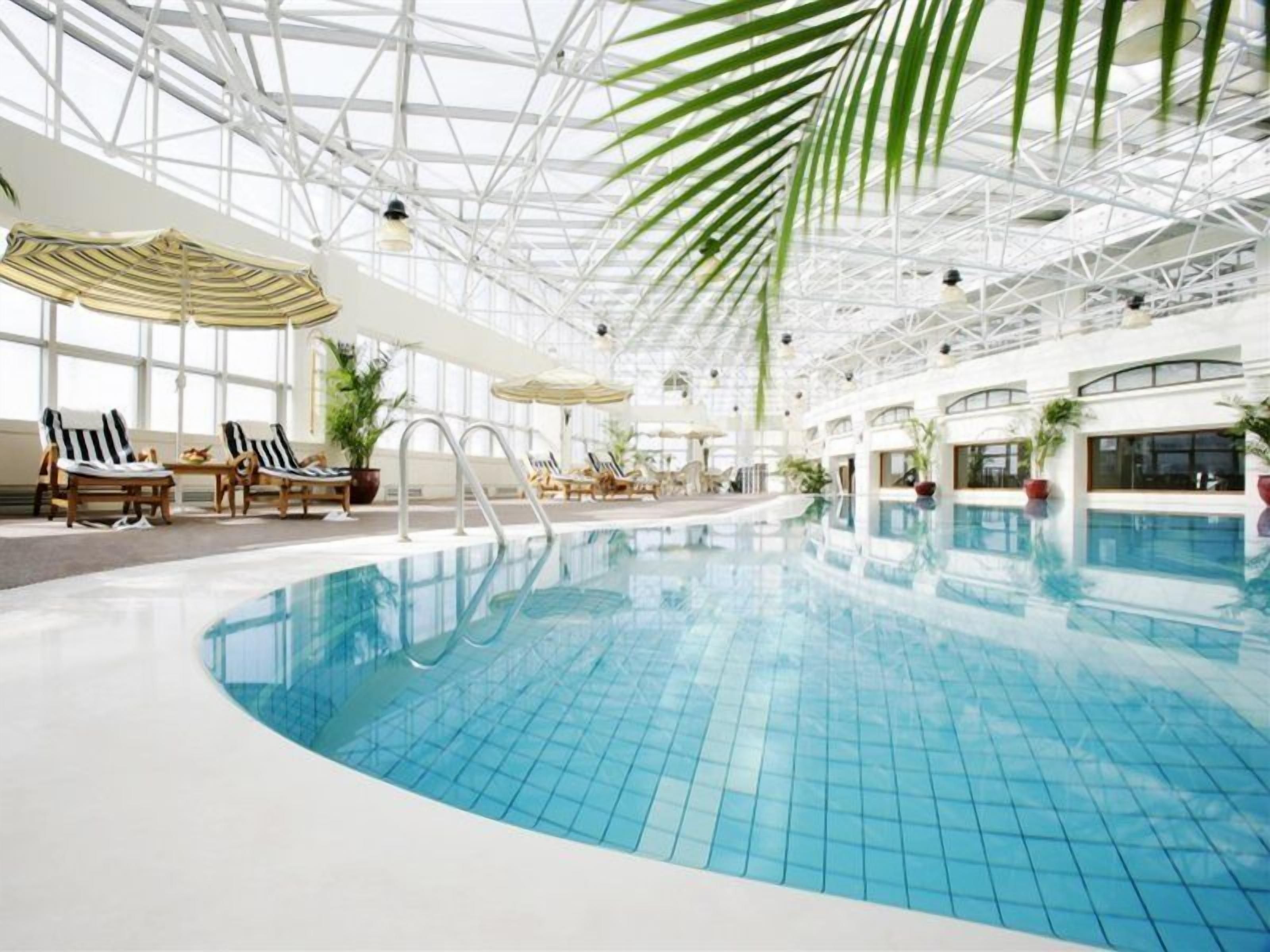 An indoor swimming pool with lounge chairs and umbrellas, surrounded by a high ceiling with a glass structure.