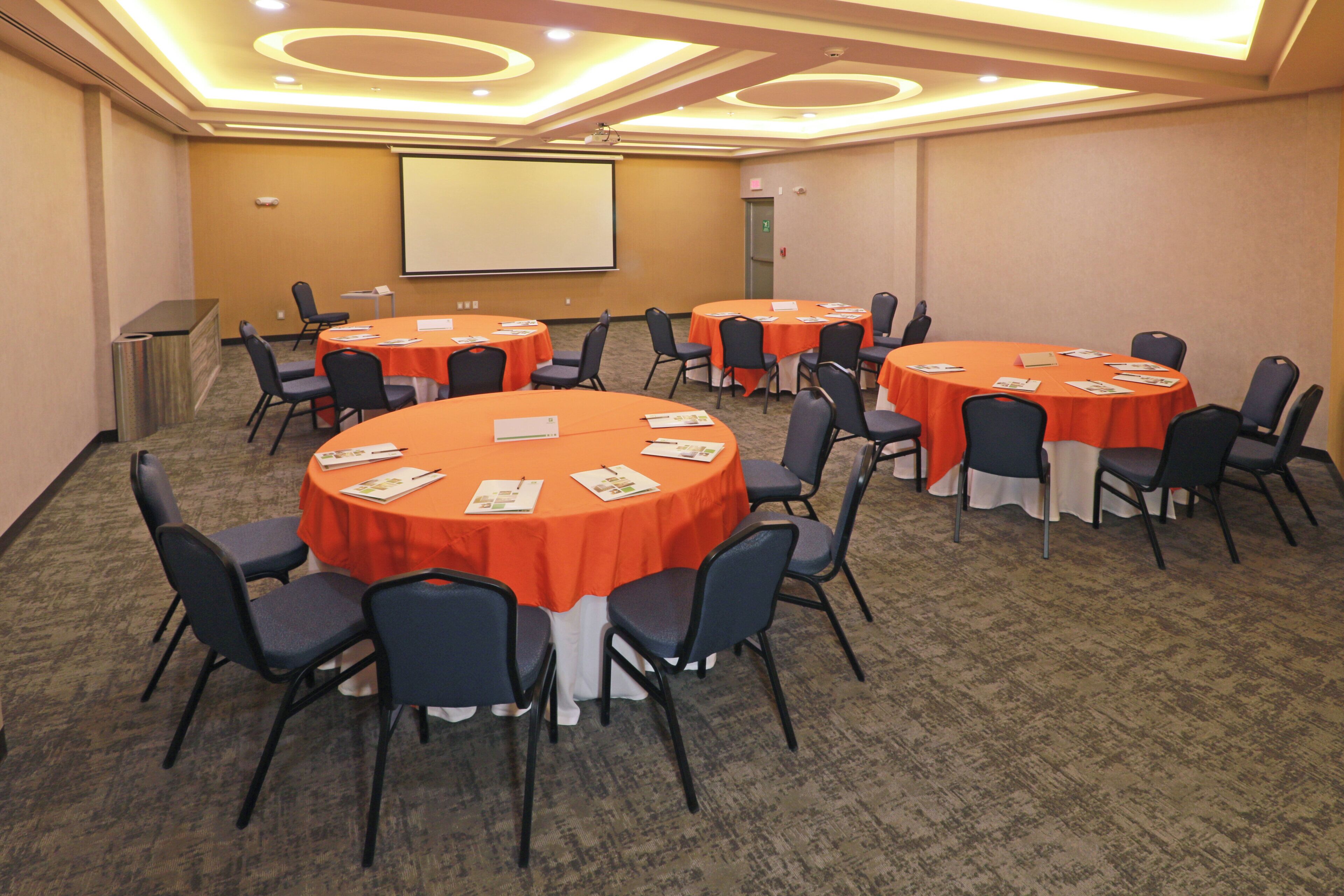 Une salle de conférence dotée de plusieurs tables rondes, chacune recouverte d’une nappe blanche et entourée de chaises.