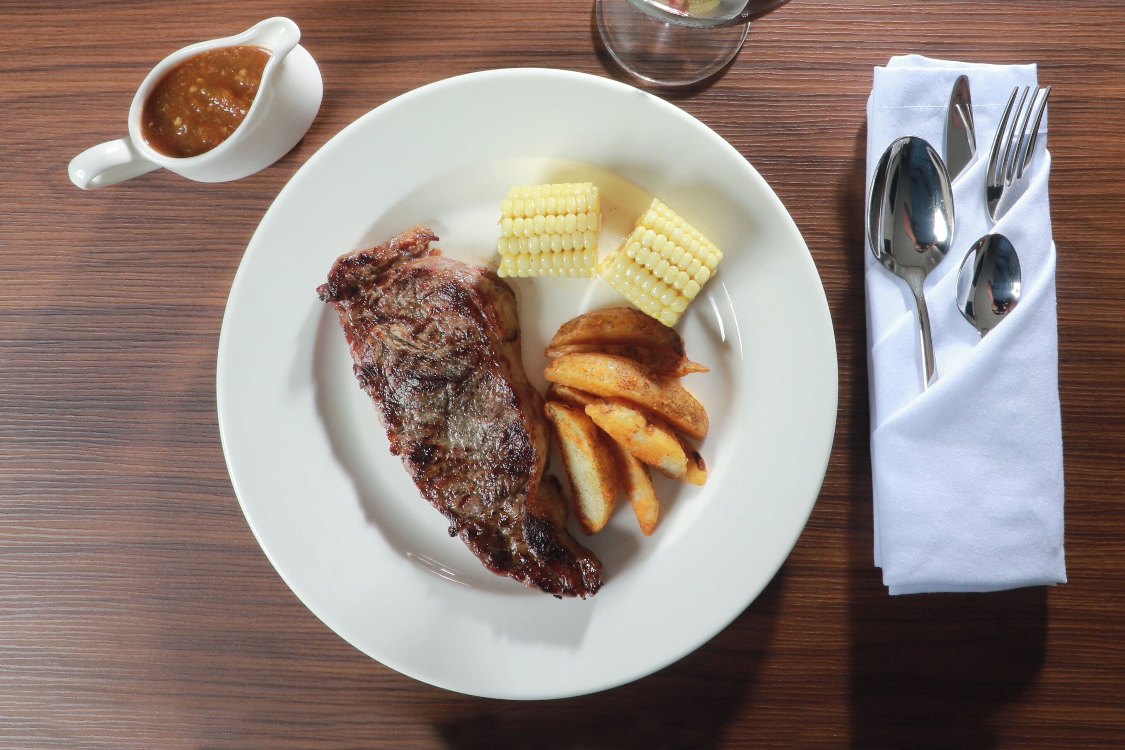 Une assiette contenant un steak, des épis de maïs et des frites, posée sur une table en bois.