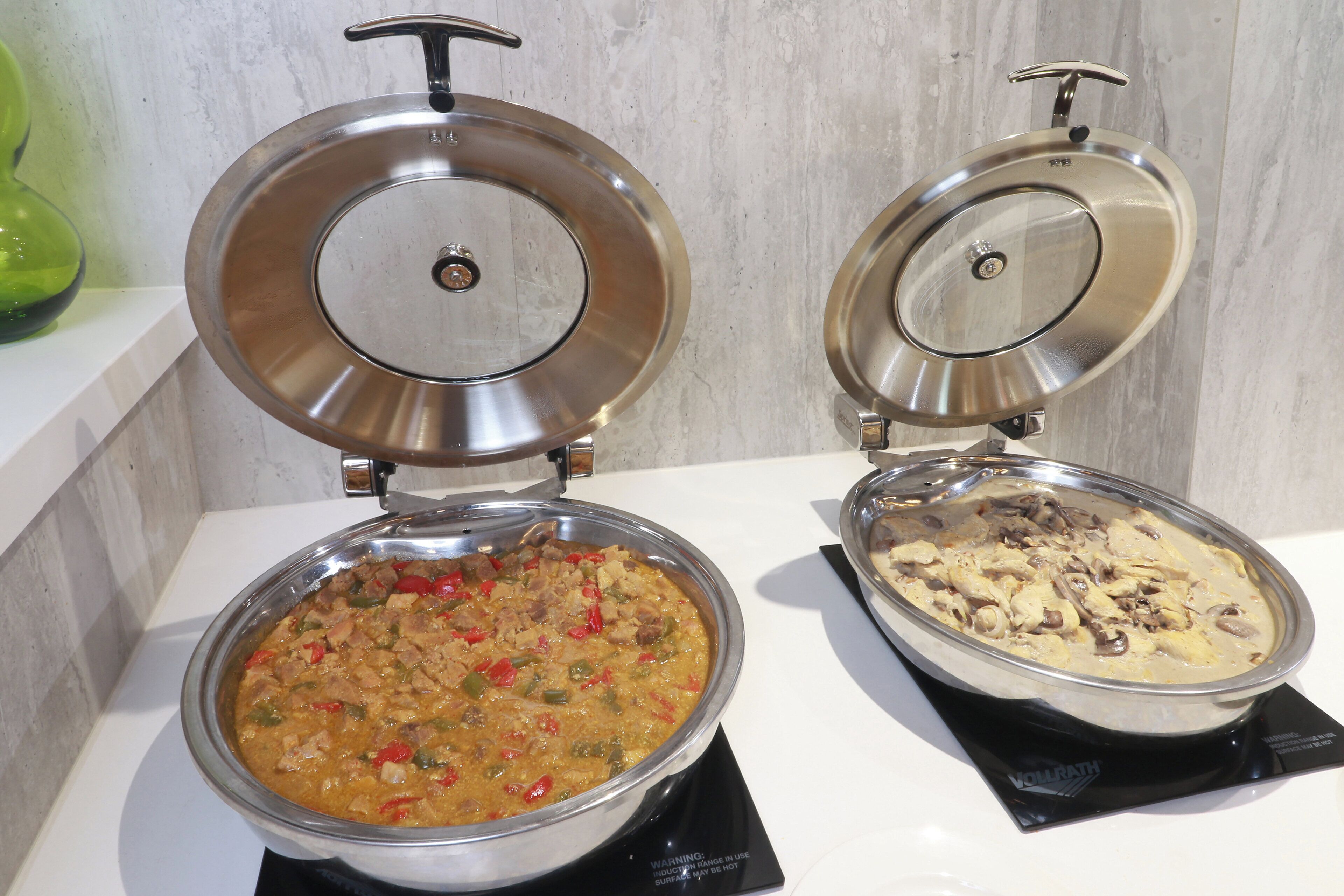 Twee roestvrijstalen voedingsdispensers met deksels, waarvan één een schotel met rood en groen groentegerecht bevat en de andere een schotel met kip in roomsaus.