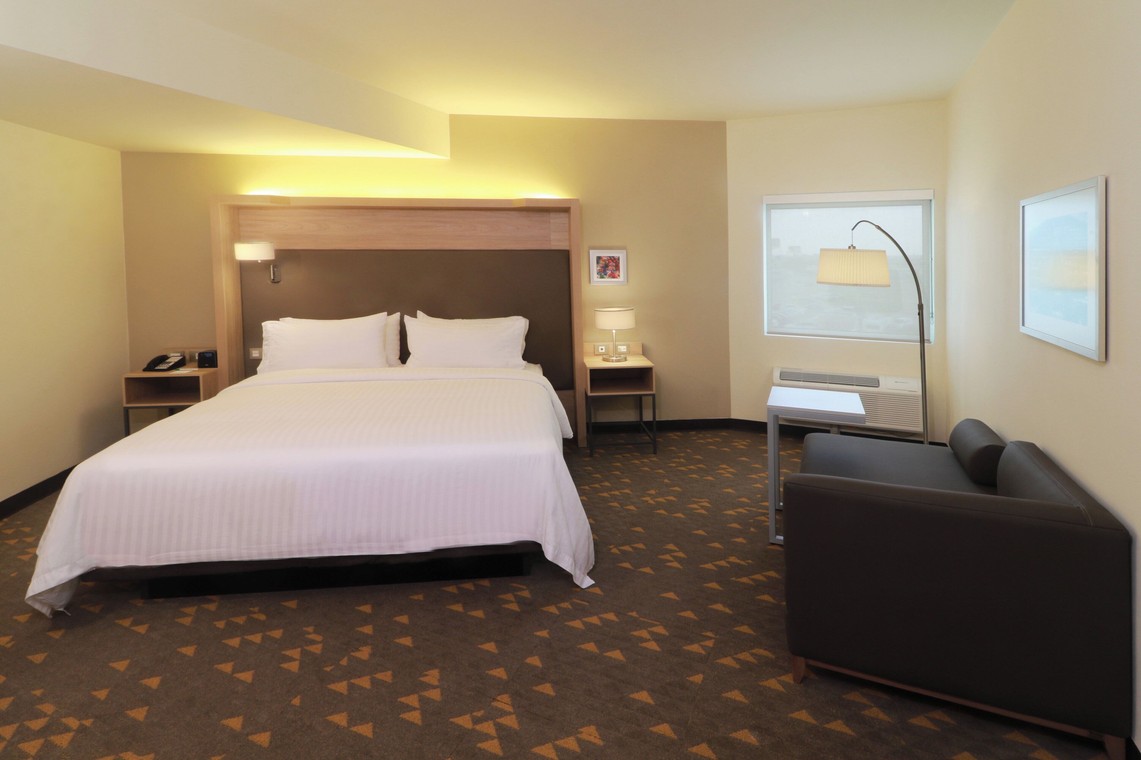 A hotel room with a large bed, bedside tables, a desk, a chair, a window, and a wall-mounted lamp.
