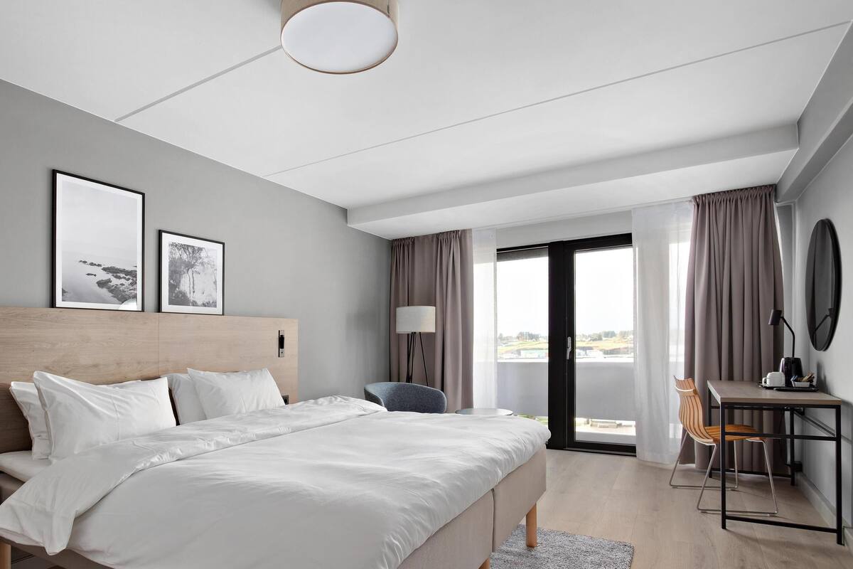 A modern hotel room with a large bed, a desk, and a chair. There are two framed pictures on the wall, a lamp, and a view of the outdoors through the window.