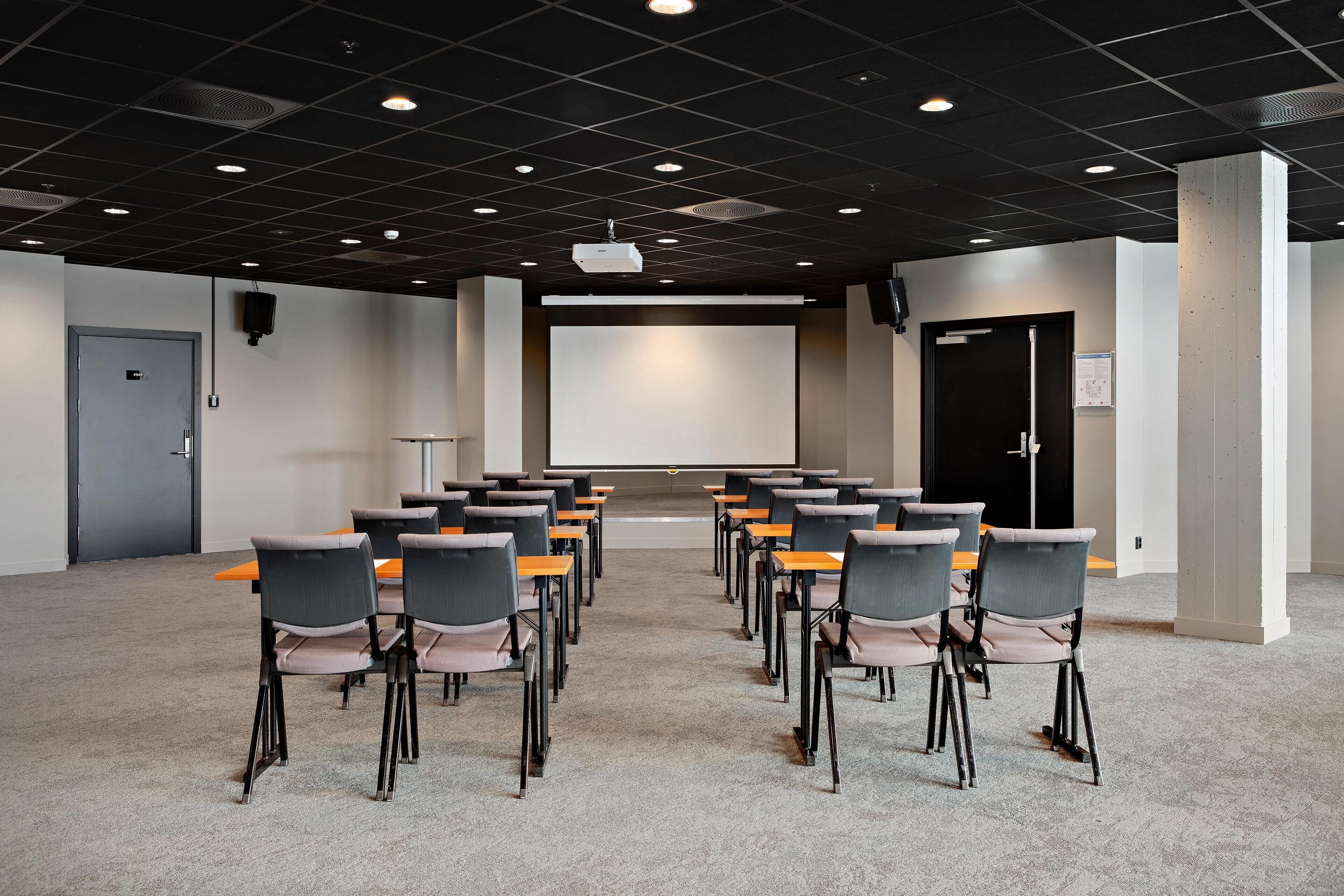 A conference room with rows of chairs facing a large screen, a projector, and a door.
