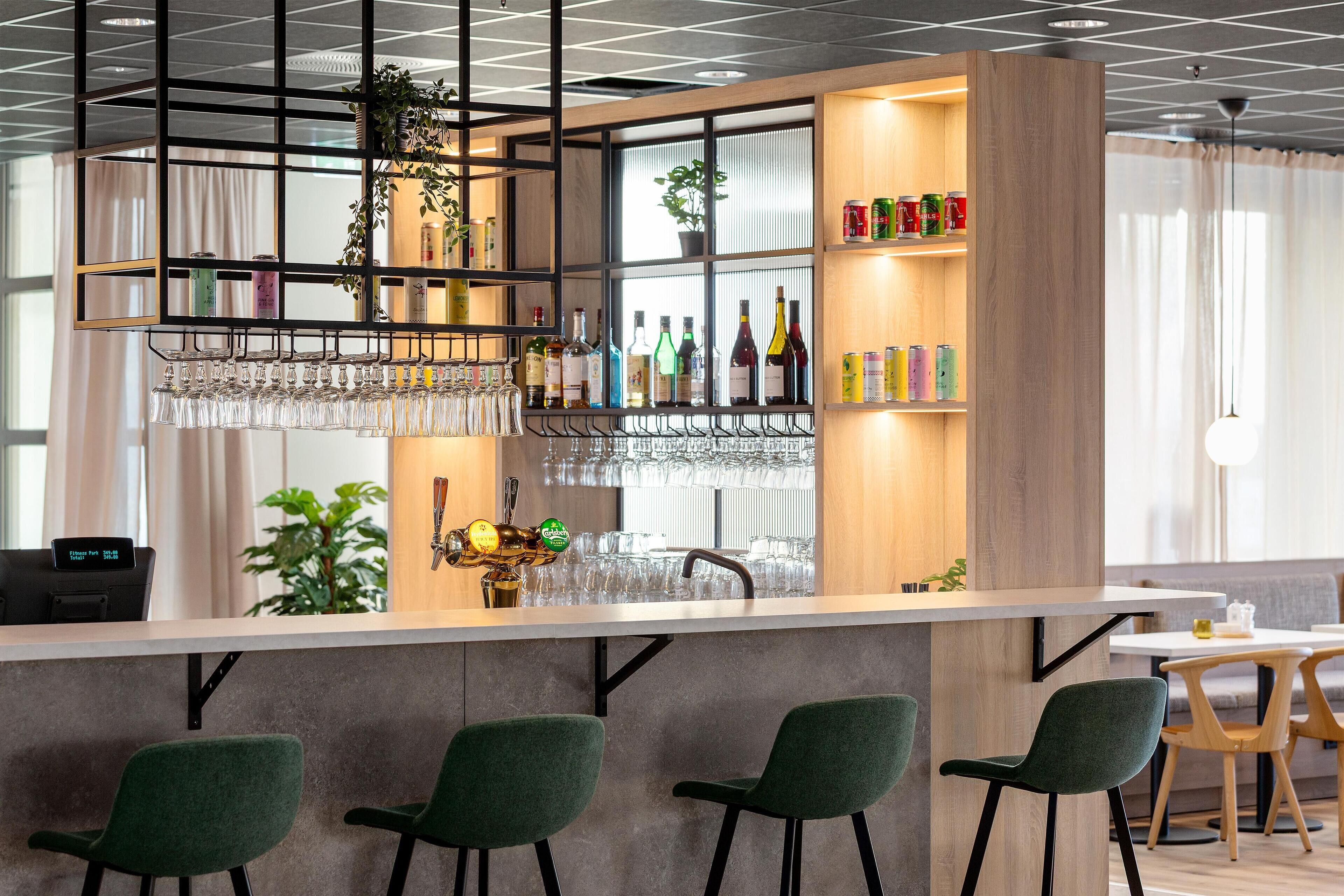 Un área de bar moderna con sillas verdes, una barra, estantes con botellas y una planta colgante.