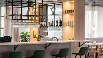 Un área de bar moderna con sillas verdes, una barra, estantes con botellas y una planta colgante.