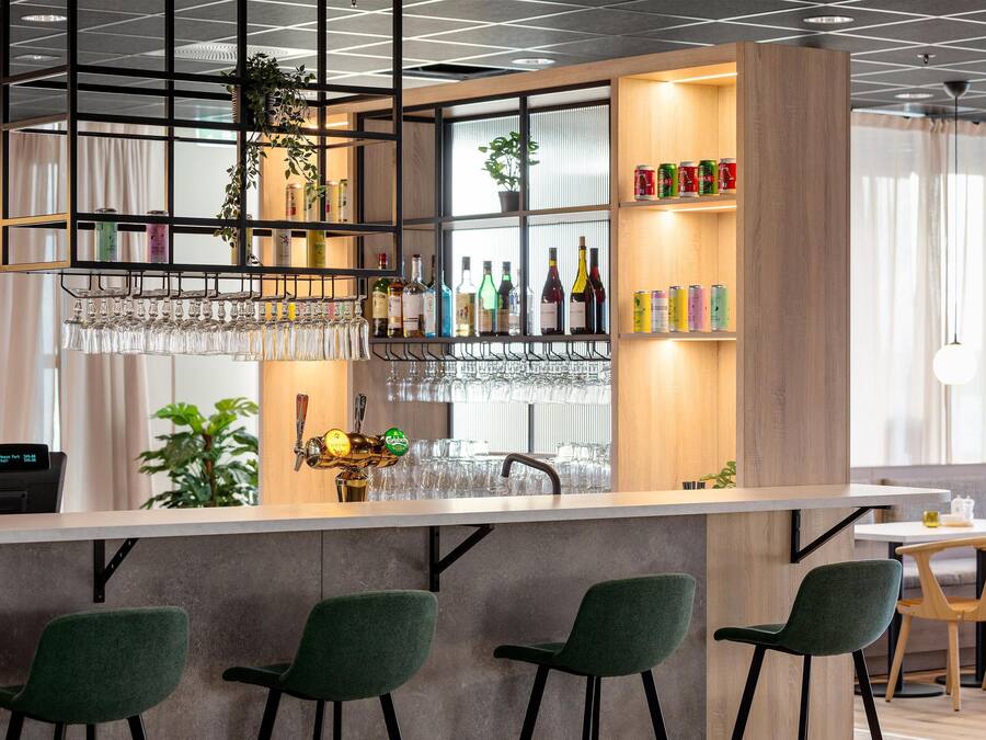 Un área de bar moderna con sillas verdes, una barra, estantes con botellas y una planta colgante.