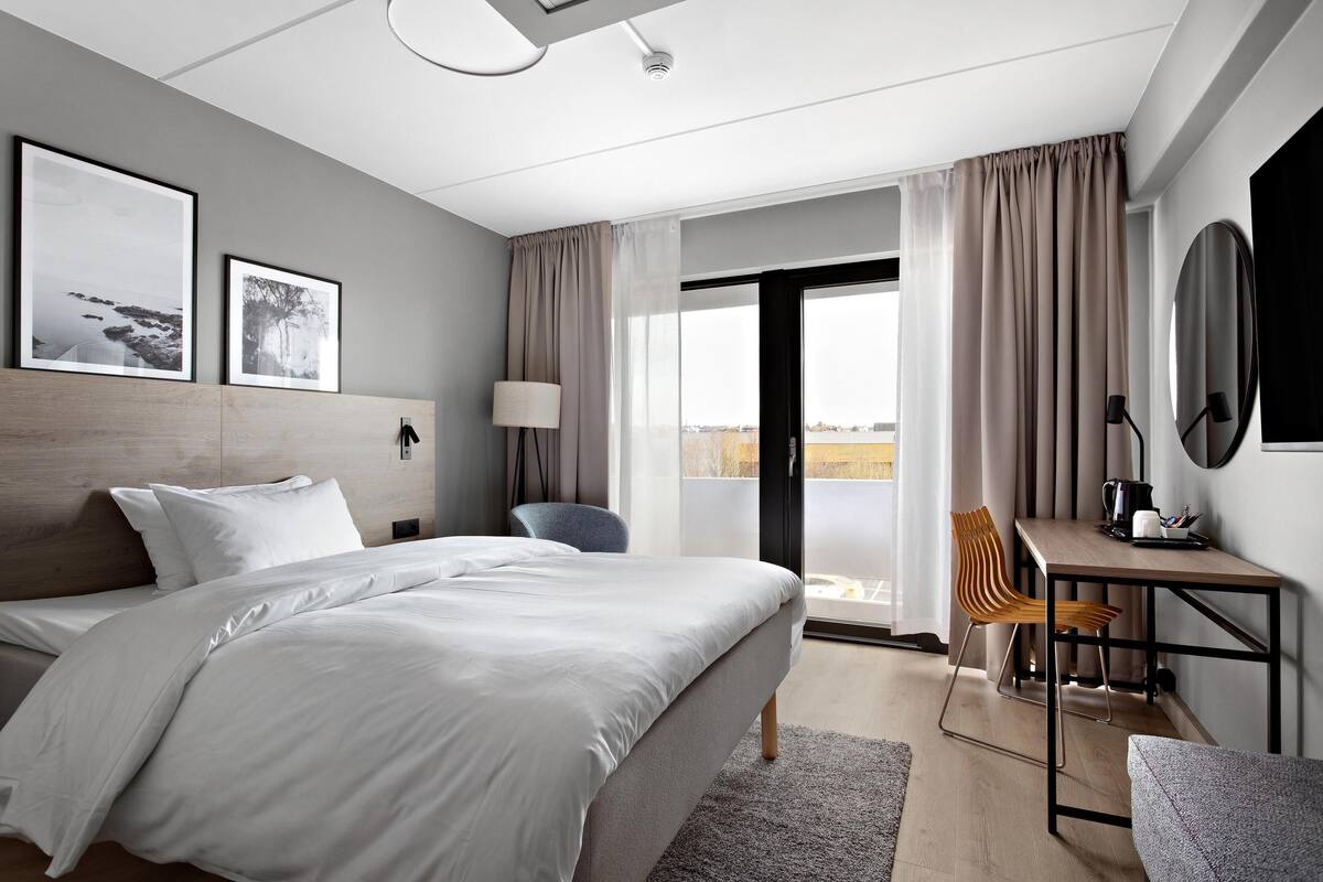 A modern hotel room with a large bed, a desk, and a chair. There are two framed pictures on the wall, a lamp, and a view of the city through the window.