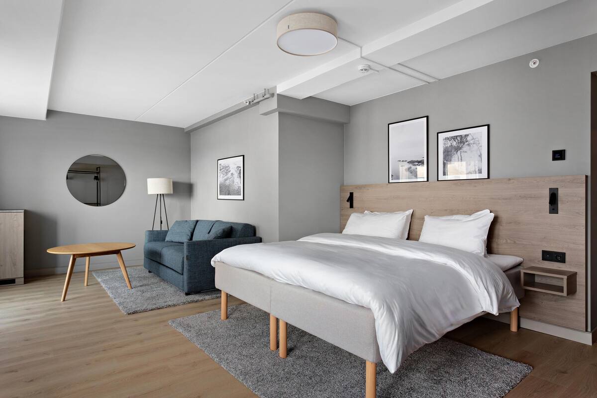 A modern bedroom with a large bed, a round mirror, a small table, a sofa, and two framed pictures on the wall.
