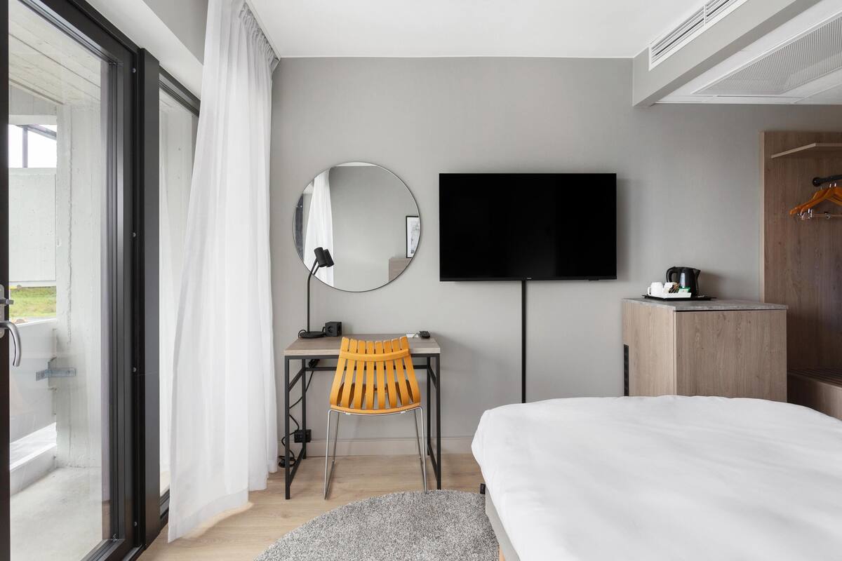 A modern hotel room with a bed, a desk with a chair, a television, and a wardrobe.