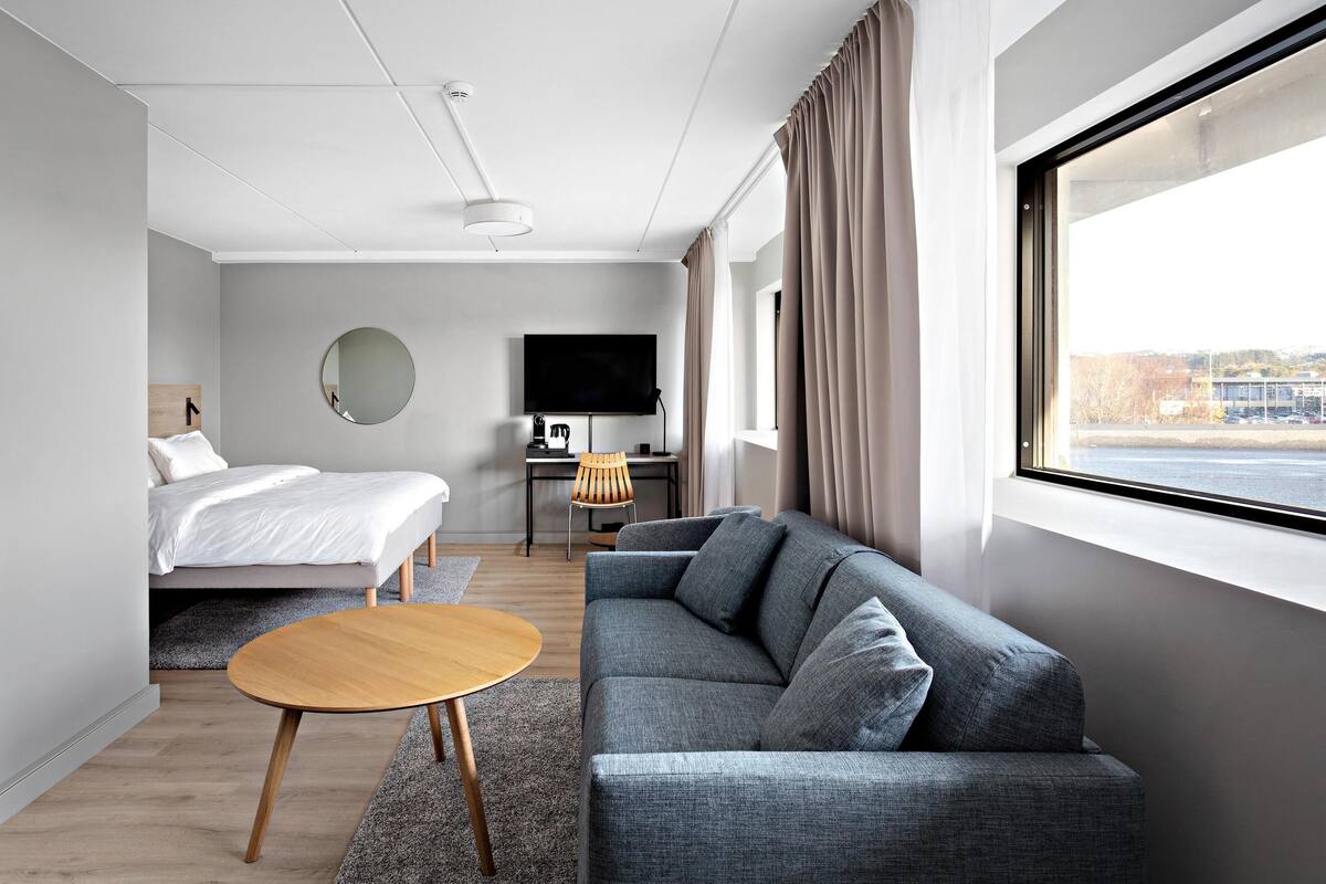 A modern hotel room with a bed, a gray sofa, a round wooden coffee table, a TV, and a large window with curtains.