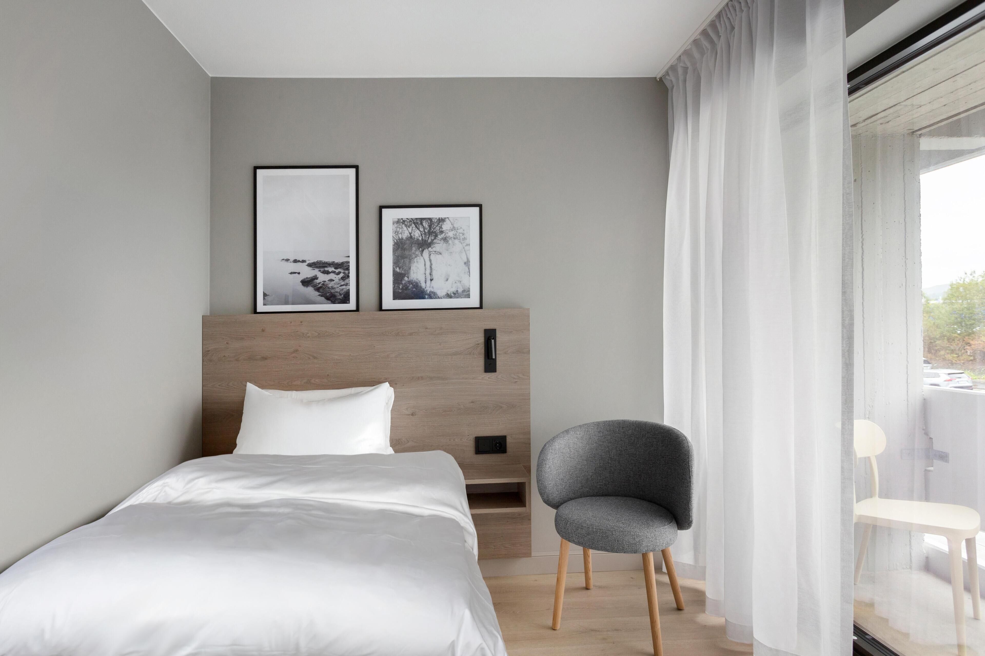 Un dormitori modern amb llit, un amb prou faena de fusta, dues fotografies amb marc, una cadira gris i una finestra amb cortines primes.