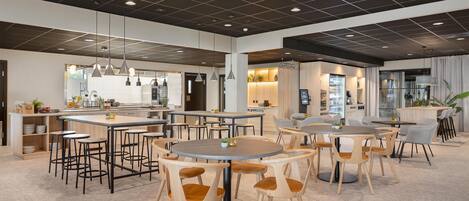 Un comedor amplio y moderno con varias mesas redondas y sillas, una cocina al fondo e iluminación empotrada en el techo.