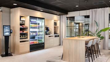 Una hall moderna di un hotel con un'isola centrale, sgabelli da bar, un grande frigorifero per bevande e una postazione informativa digitale.