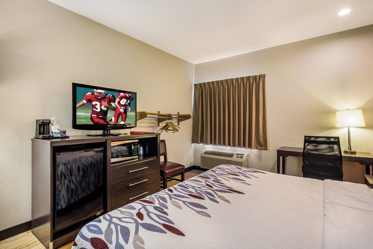 A hotel room with a bed, a television, a desk with a chair, a window with curtains, and an air conditioning unit.