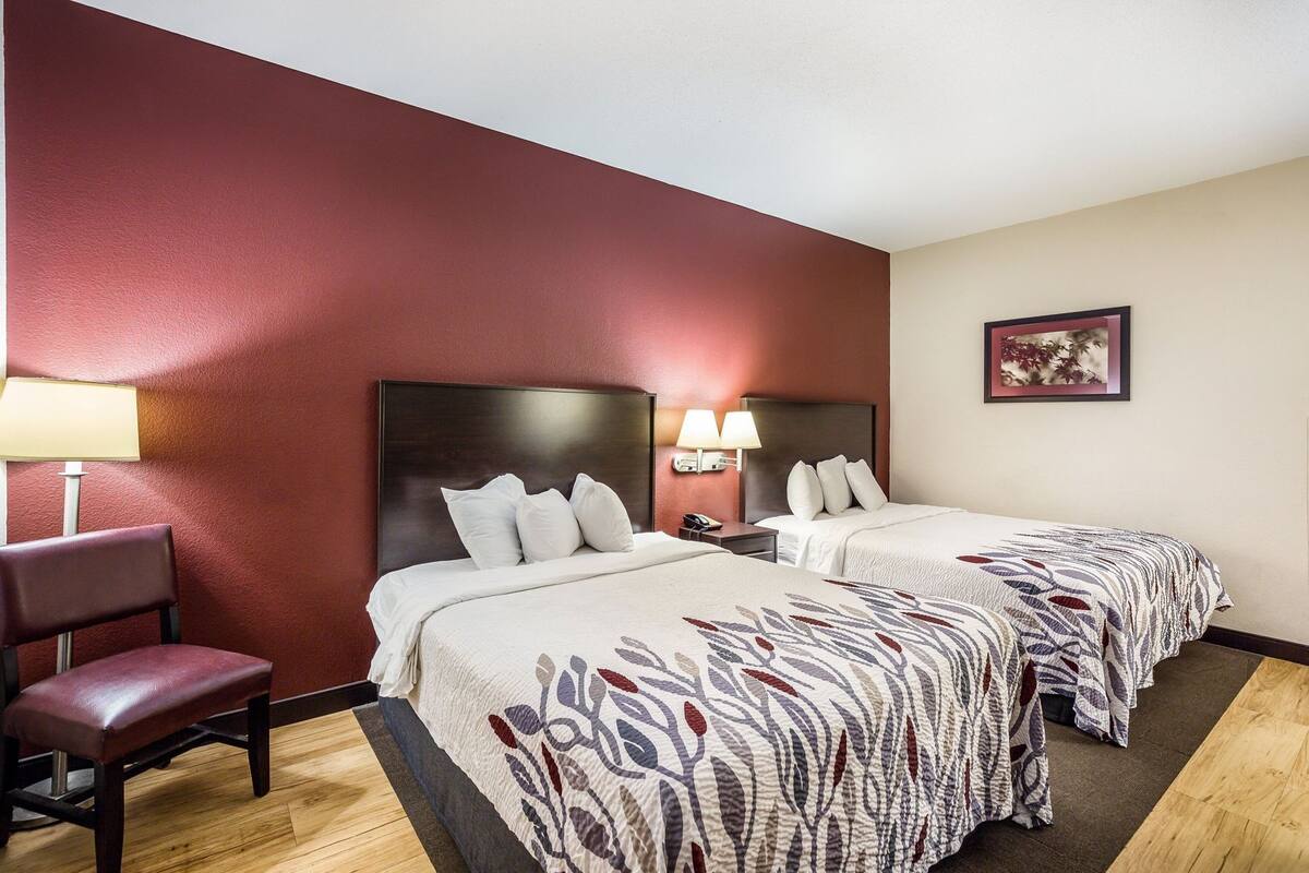 A hotel room with two beds, a chair, a nightstand, a lamp, and a framed picture on the wall.