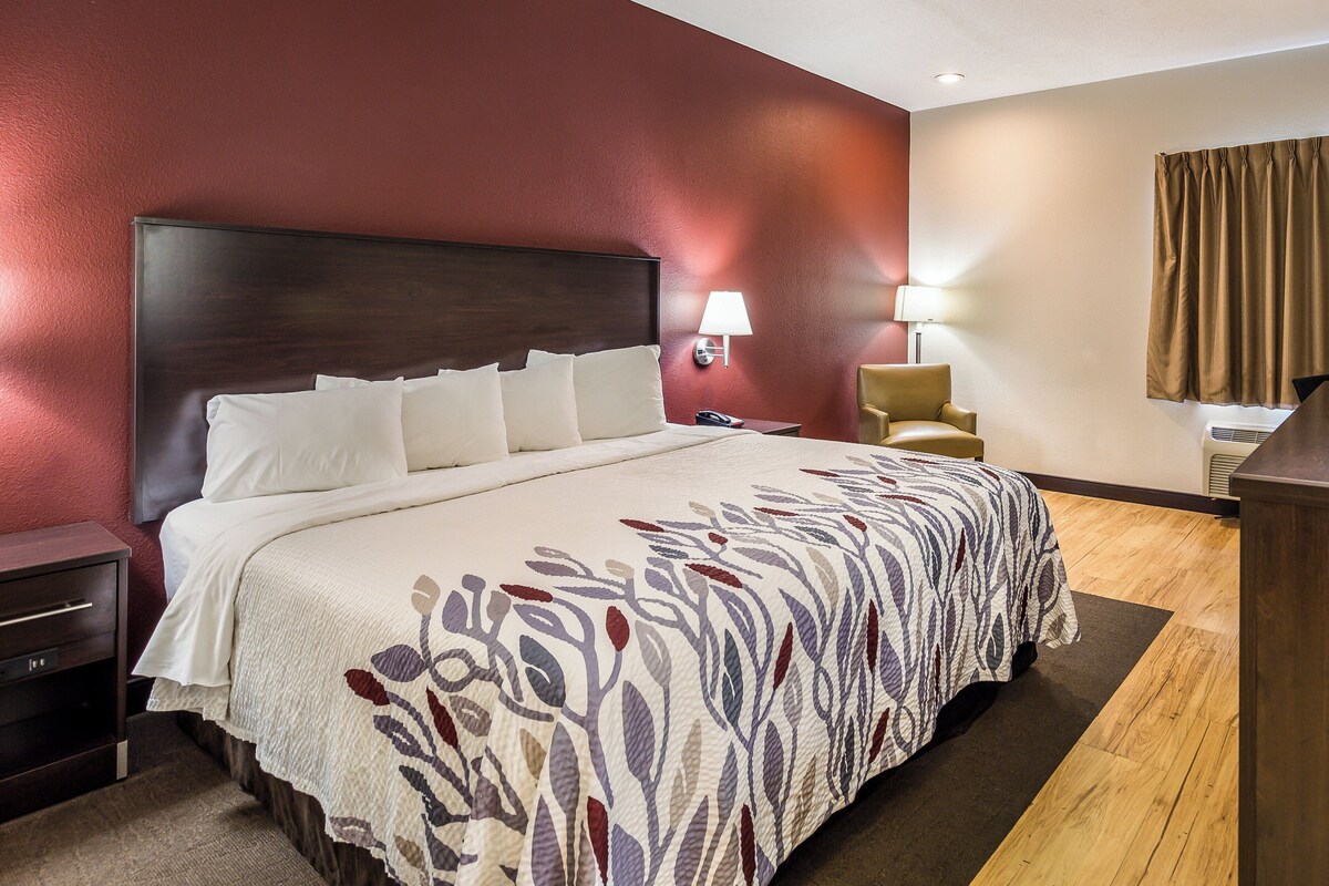 A hotel room with a large bed, a nightstand, a chair, and a desk with a lamp.