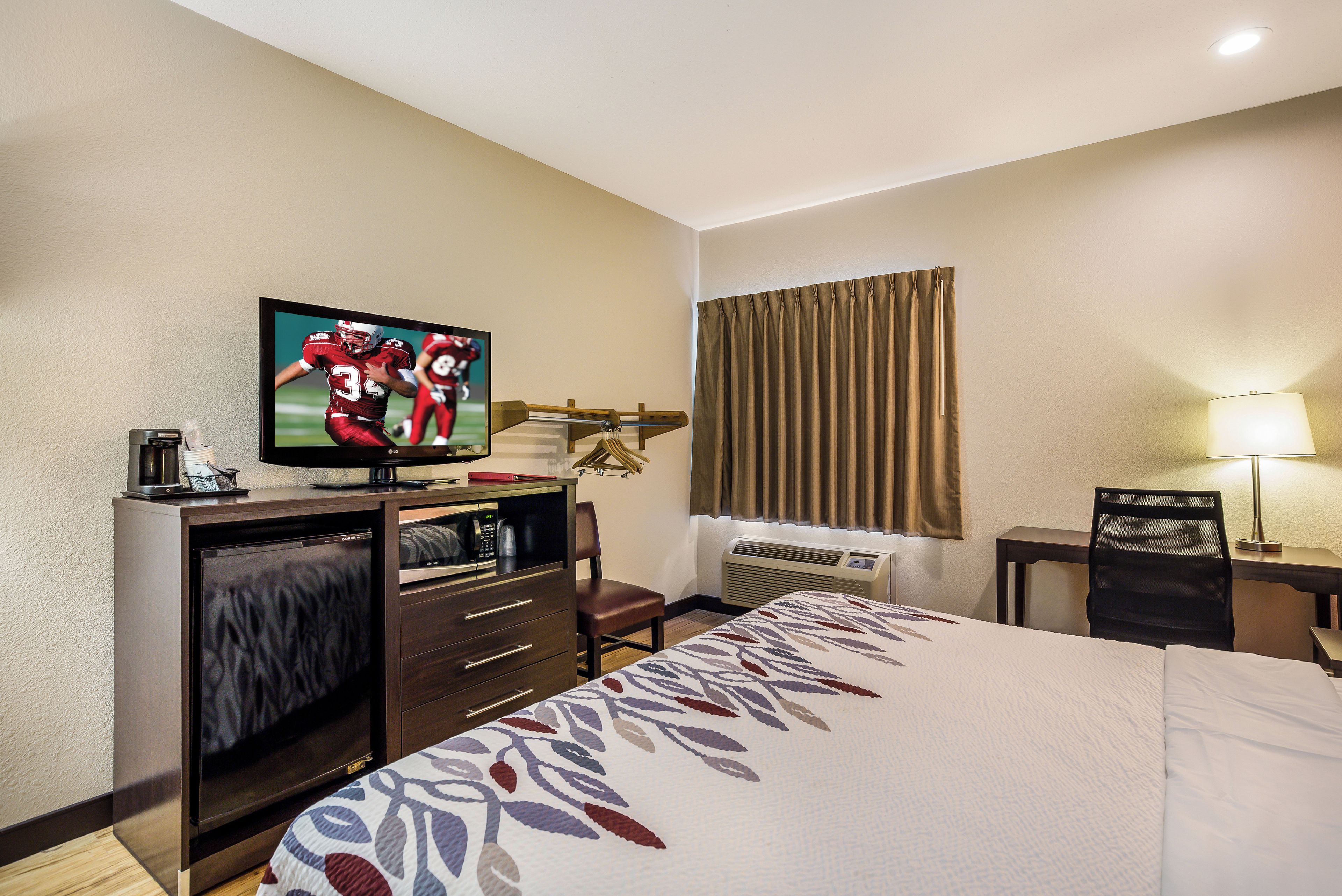 A hotel room with a bed, a television, a desk with a chair, a window with curtains, and an air conditioning unit.