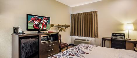 A hotel room with a bed, a television, a desk with a chair, a window with curtains, and an air conditioning unit.