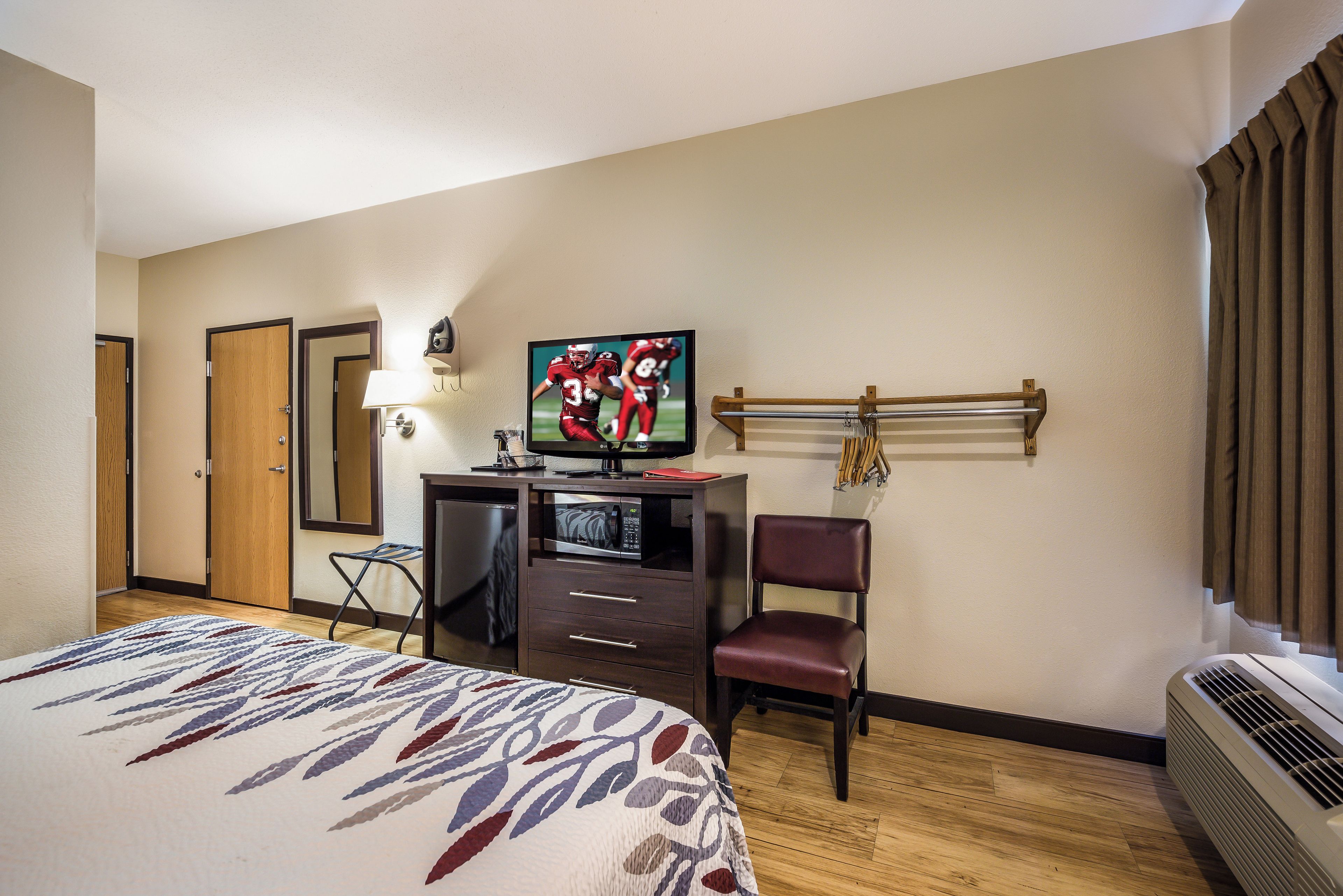 Una habitación de hotel con cama, escritorio con televisor, silla, lámpara de escritorio y un perchero en la pared con llaves.