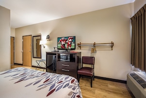 Una habitación de hotel con cama, escritorio con televisor, silla, lámpara de escritorio y un perchero en la pared con llaves.