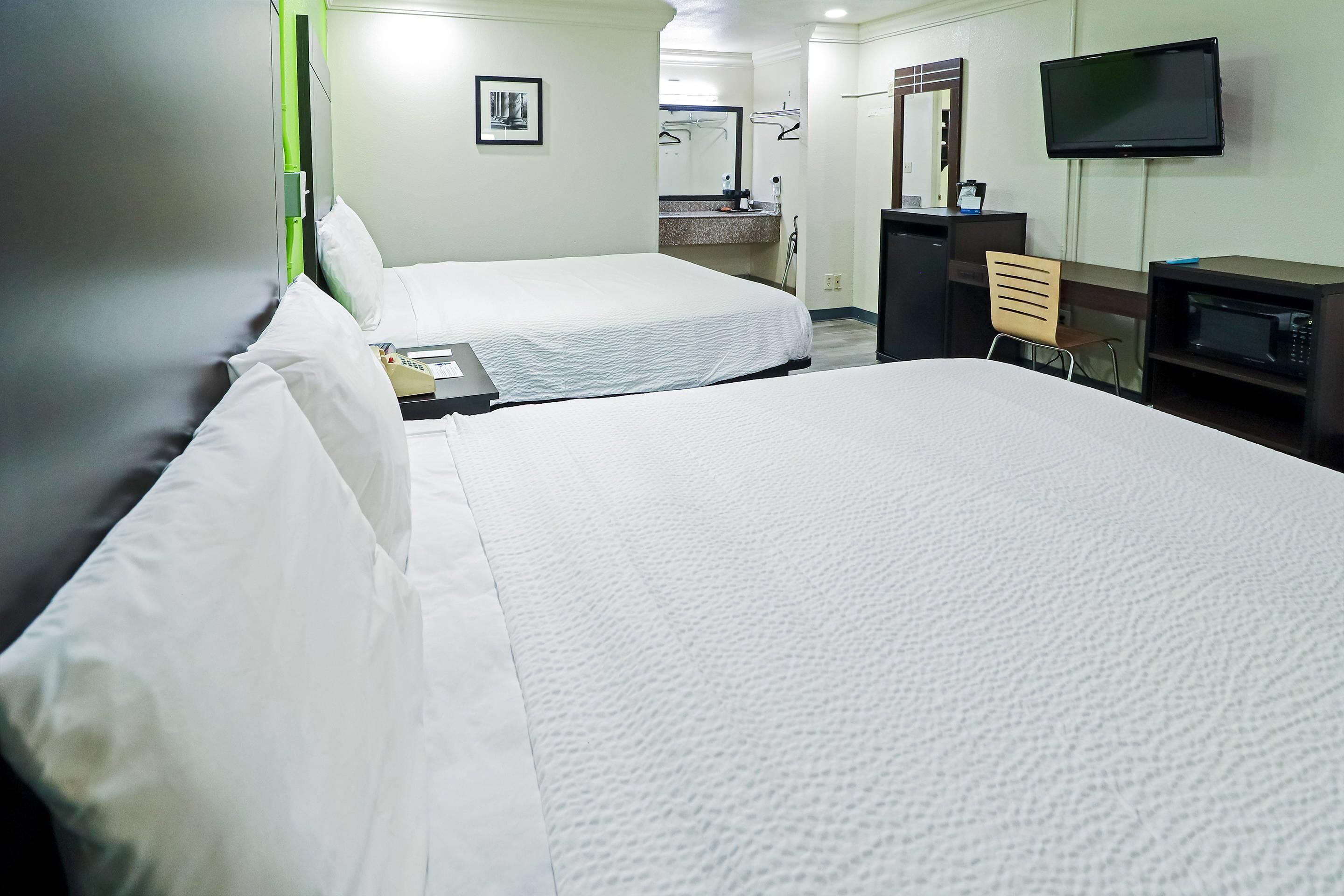 A hotel room with two beds, a desk with a chair, a television, a microwave, and a bathroom with a sink and mirror.