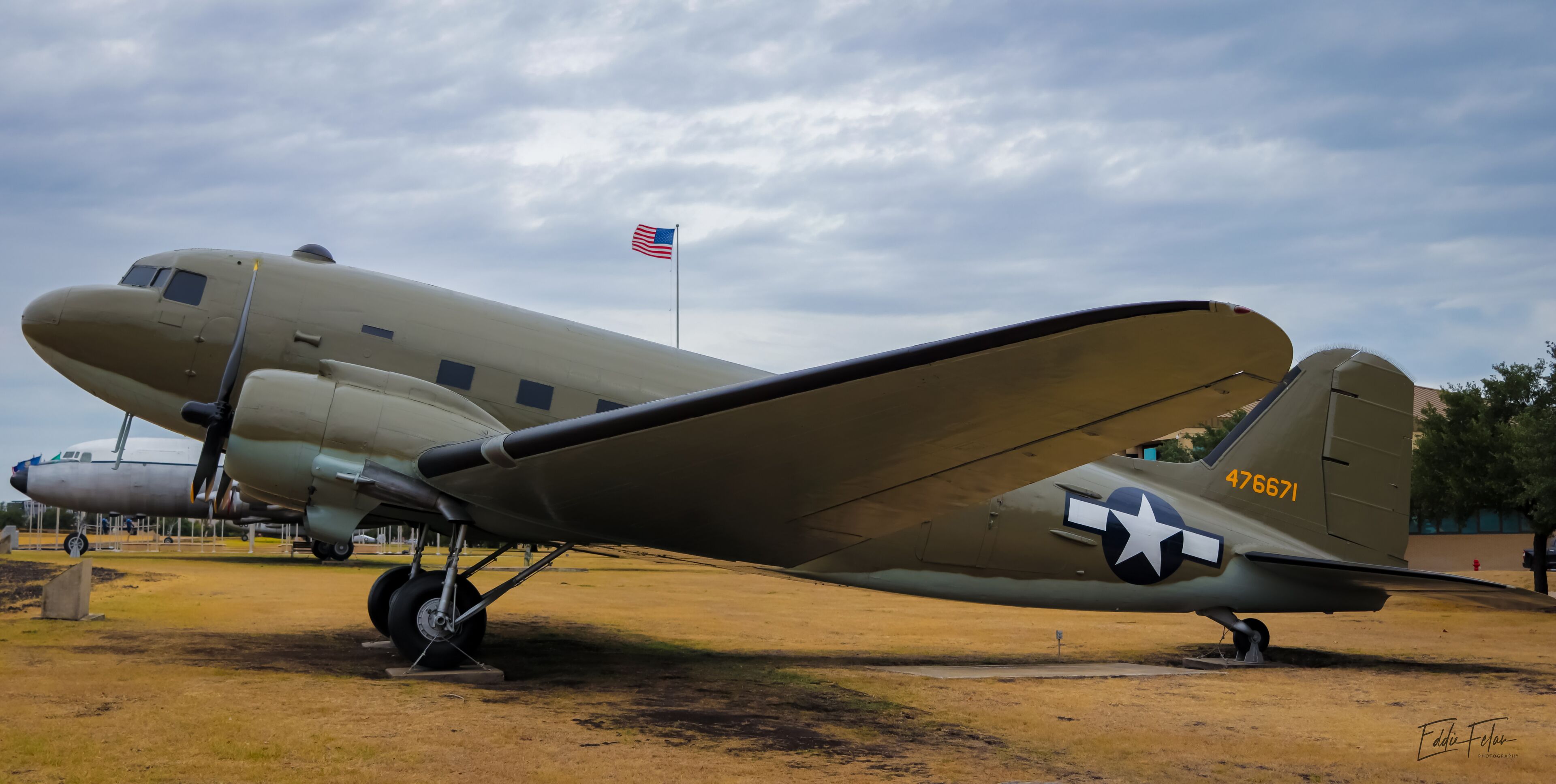 A vintage military aircraft with a star insignia and the number 476671 on its tail, parked on a grassy field with a flag in the background.
