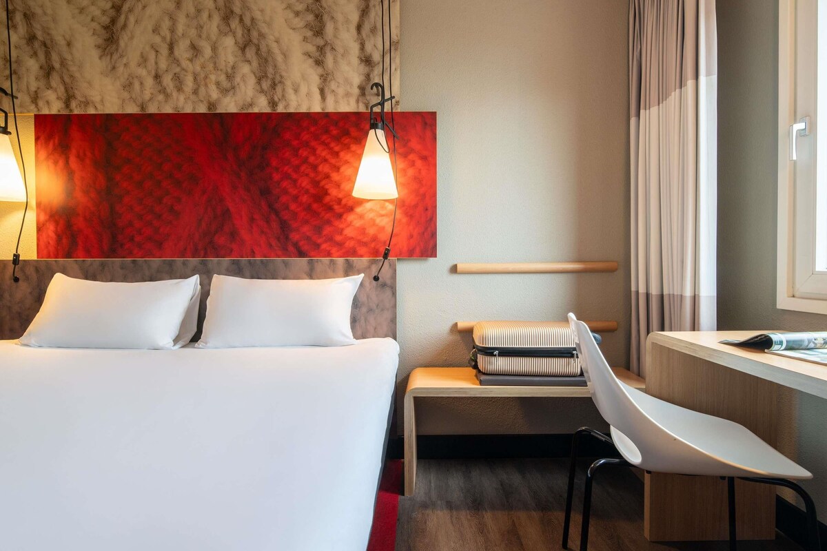 A hotel room with a bed, a desk, a chair, a window with curtains, and a red textured wall.