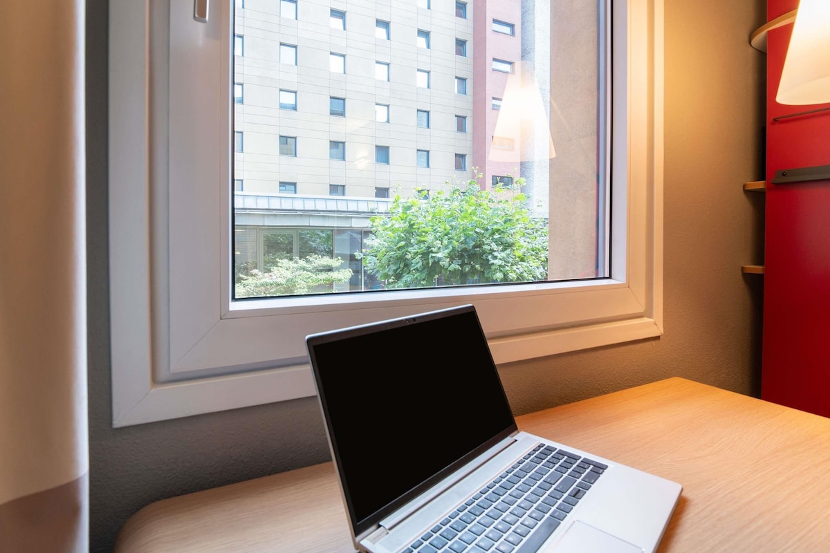 A laptop on a table next to a window with a view of a building and some greenery.