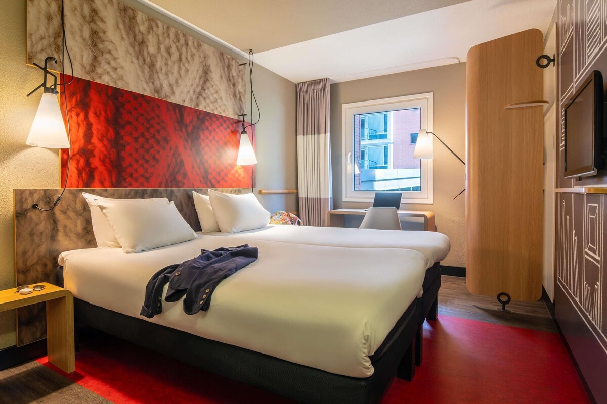 A hotel room with a bed, bedside tables, a desk, a TV, and a window with a view of buildings.