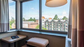 Habitación de hotel con un ventanal grande, una mesita, una maleta y una lámpara.