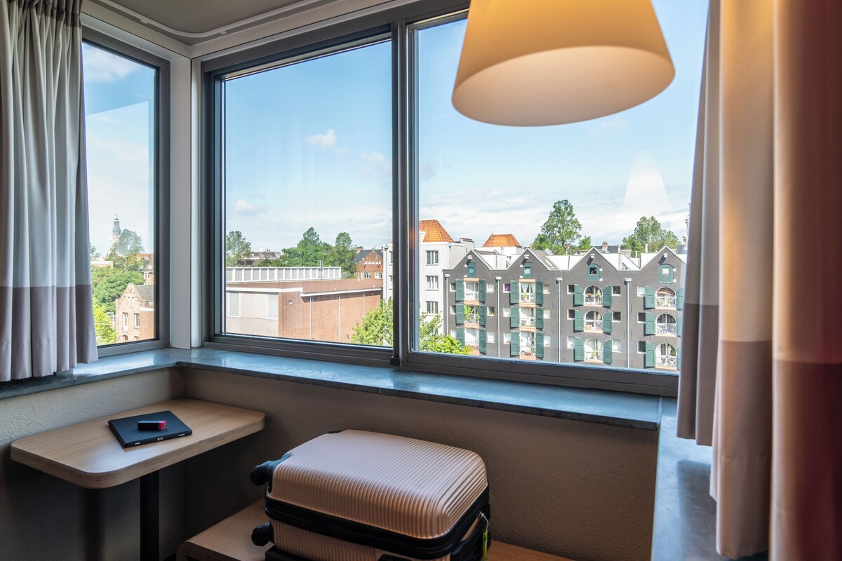 A hotel room with a large window, a small table, a suitcase, and a lamp.