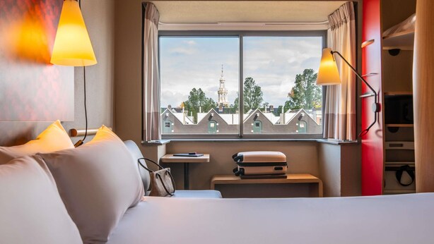 Habitación de hotel con cama, ventana que ofrece vistas a edificios, una mesita pequeña y una maleta.