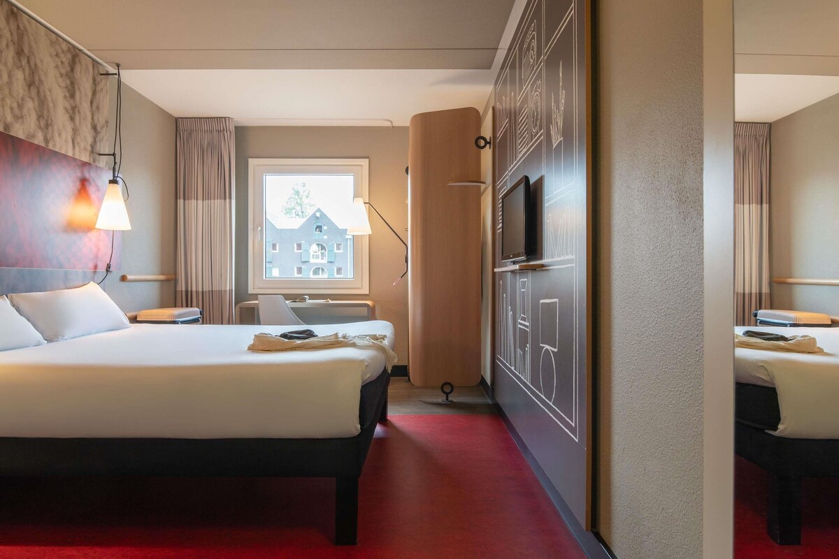 A hotel room with a bed, a desk, a TV, and a window with a view of a building.