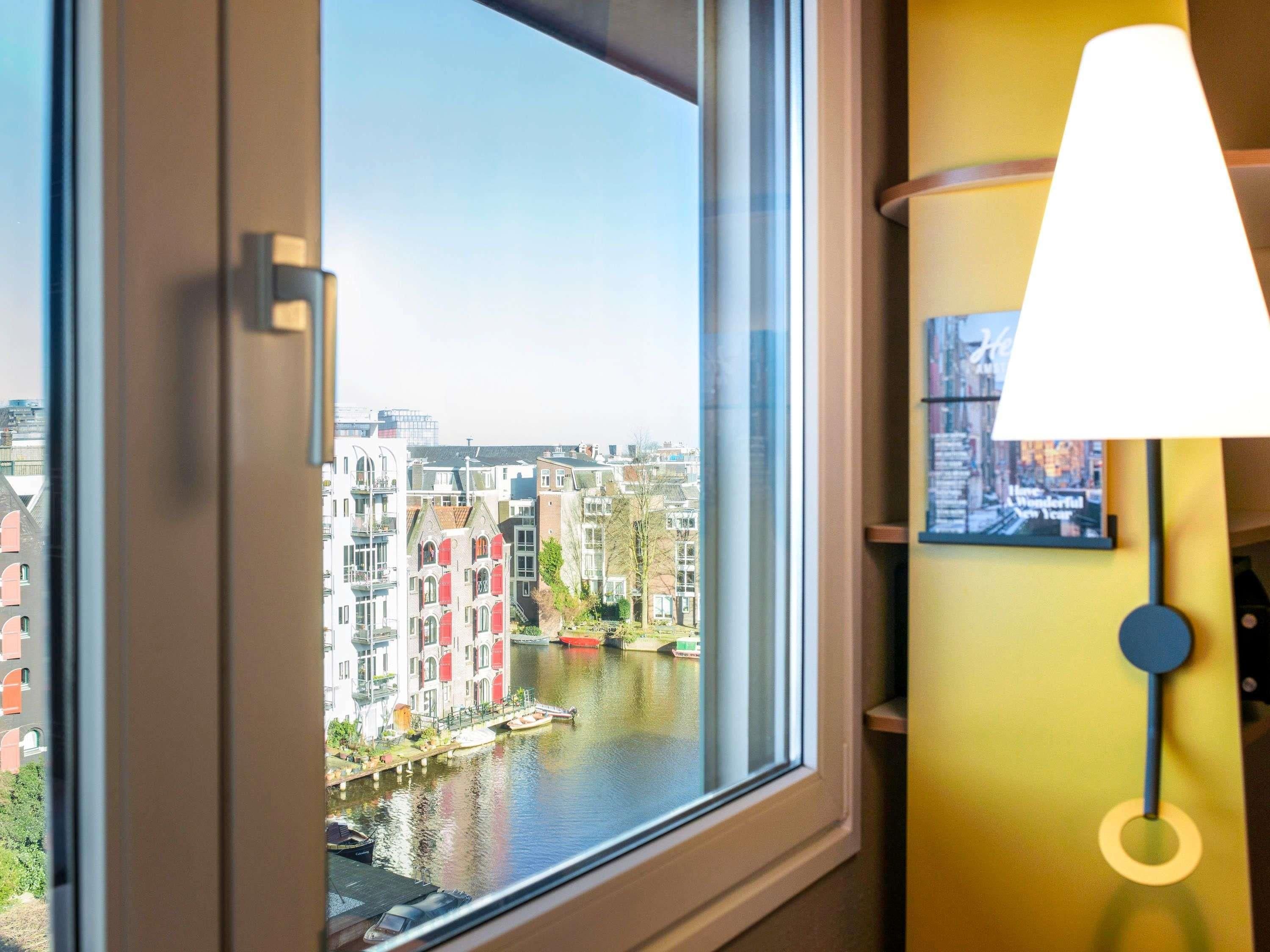 Een raam met uitzicht op een gracht en gebouwen, een lamp en een ingelijst foto aan de muur.