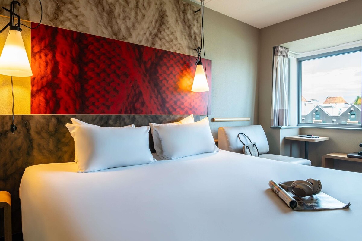 A hotel room with a large bed, two bedside lamps, a window with a view of buildings, and a small table with a magazine and headphones.