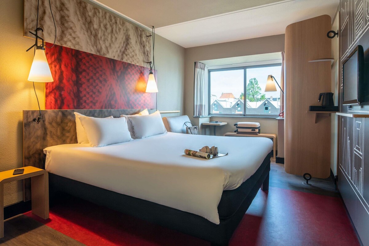 A hotel room with a large bed, bedside tables, a TV, a window with a view, and a red carpet.