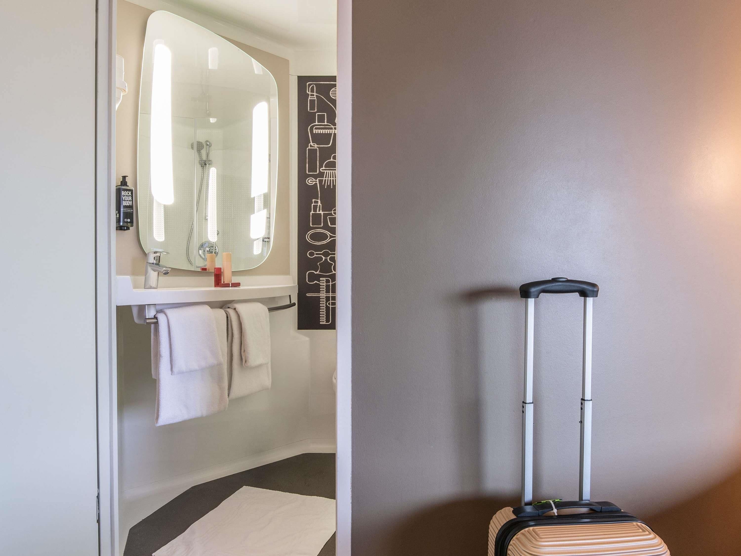 A bathroom with a large mirror, a sink, and a towel rack. There is a suitcase against the wall.